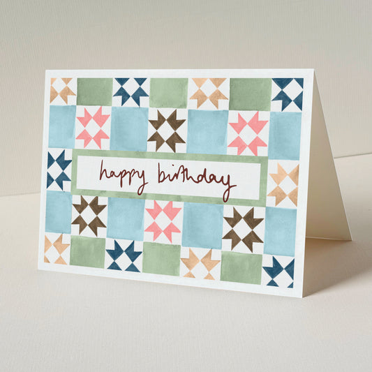 A folded greeting card standing upright on a light beige surface. The front of the card has a vintage tile design in blue, green, pink, brown, and navy, with a white centre panel and the words 'happy birthday'.