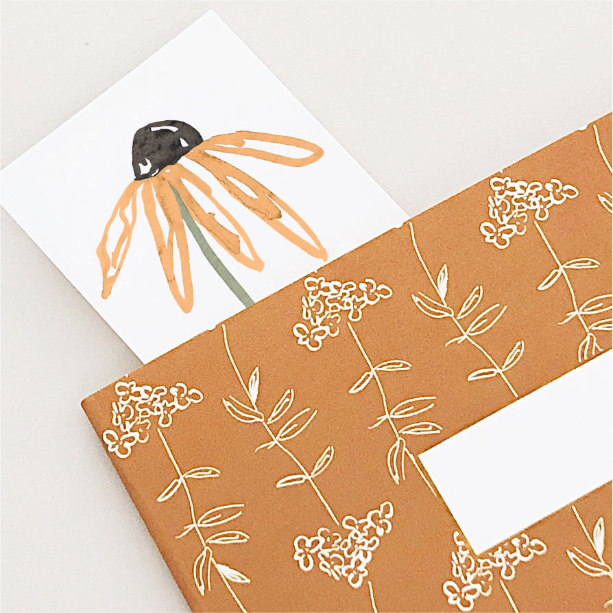 A close-up of an orange daisy bookmark inside a rust coloured floral notebook.