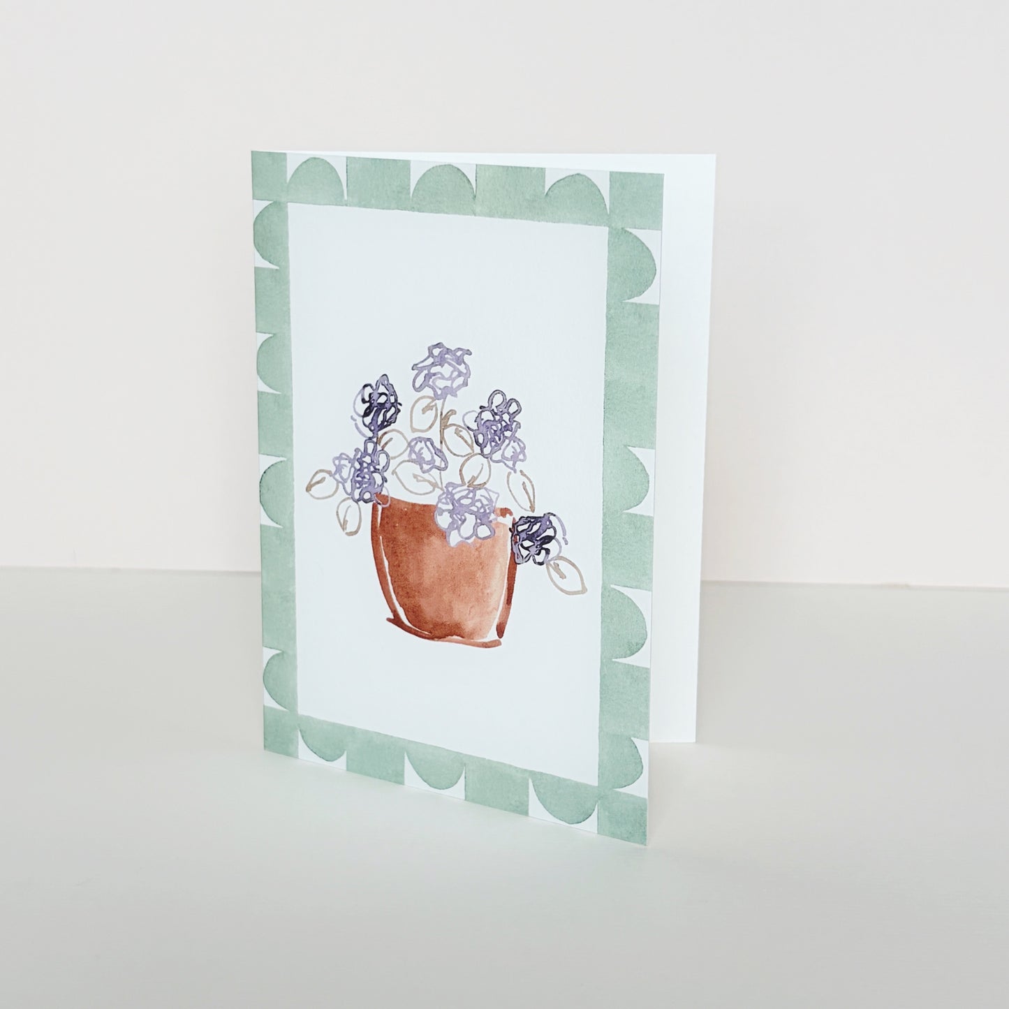 A mini greeting card with a geometric green border and a central ink illustration of a terracotta pot with purple flowers, standing upright on a light beige surface against a cream background.