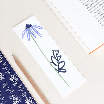 A rectangular bookmark with a hand-drawn blue daisy and green stem, placed on a cream desk with an open book, pencil, and patterned notebook nearby.