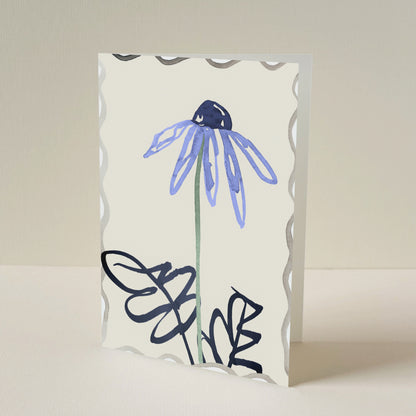 A greeting card with a scalloped  border, featuring a hand-drawn blue flower and dark navy hand drawn leaves on a warm cream background, standing upright against a neutral beige backdrop.