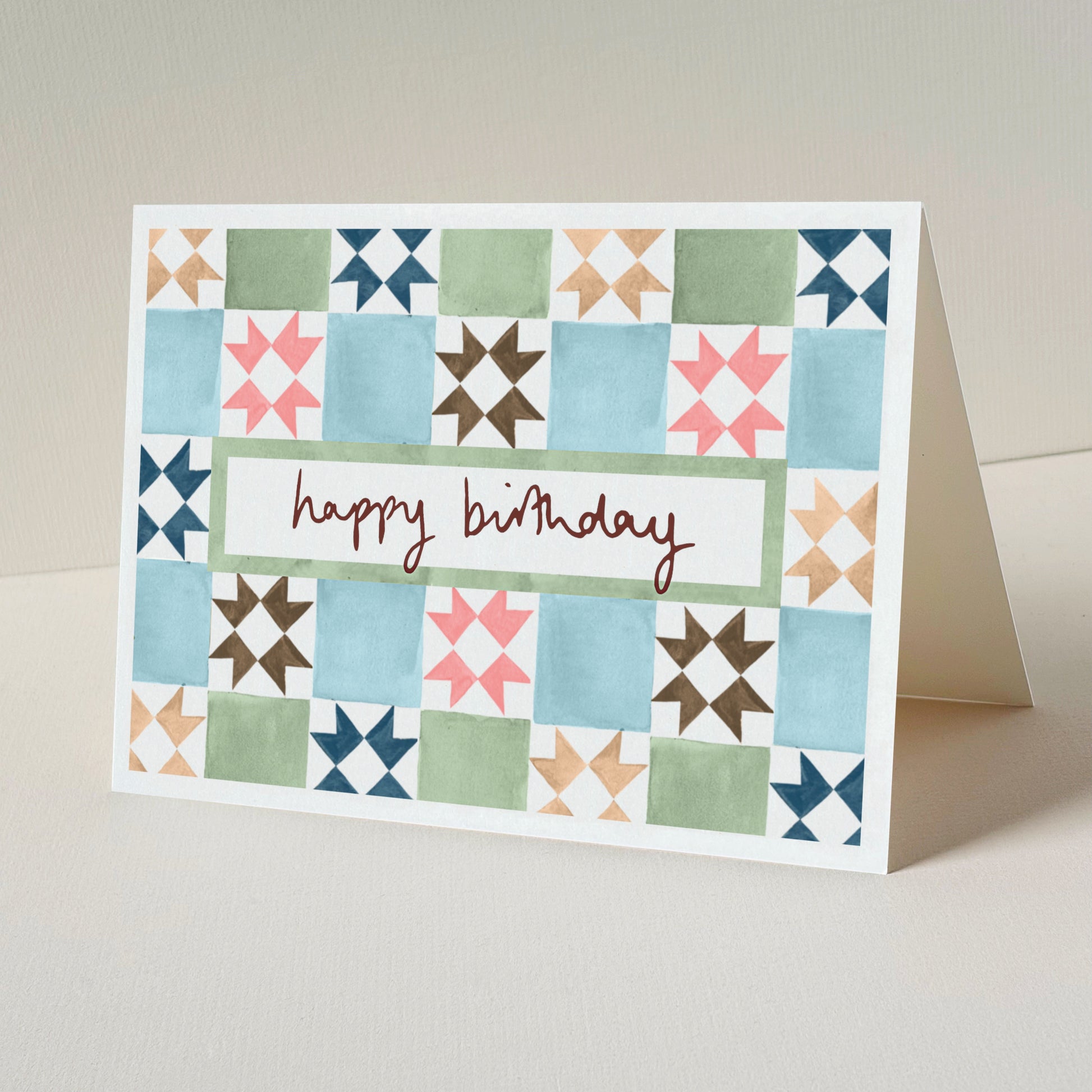 A folded greeting card standing upright on a light beige surface. The front of the card has a vintage tile design in blue, green, pink, brown, and navy, with a white centre panel and the words 'happy birthday'.