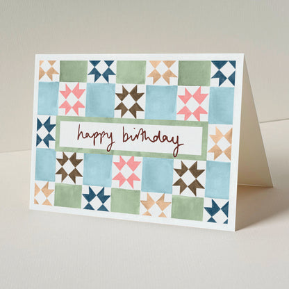 A folded greeting card standing upright on a light beige surface. The front of the card has a vintage tile design in blue, green, pink, brown, and navy, with a white centre panel and the words 'happy birthday'.