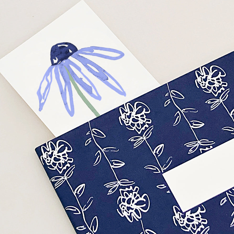 A close-up of a blue daisy bookmark inside a navy floral notebook.
