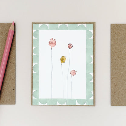 A mini greeting card with a floral bud design, displayed on a light beige background next to a pink pencil and kraft paper envelopes.