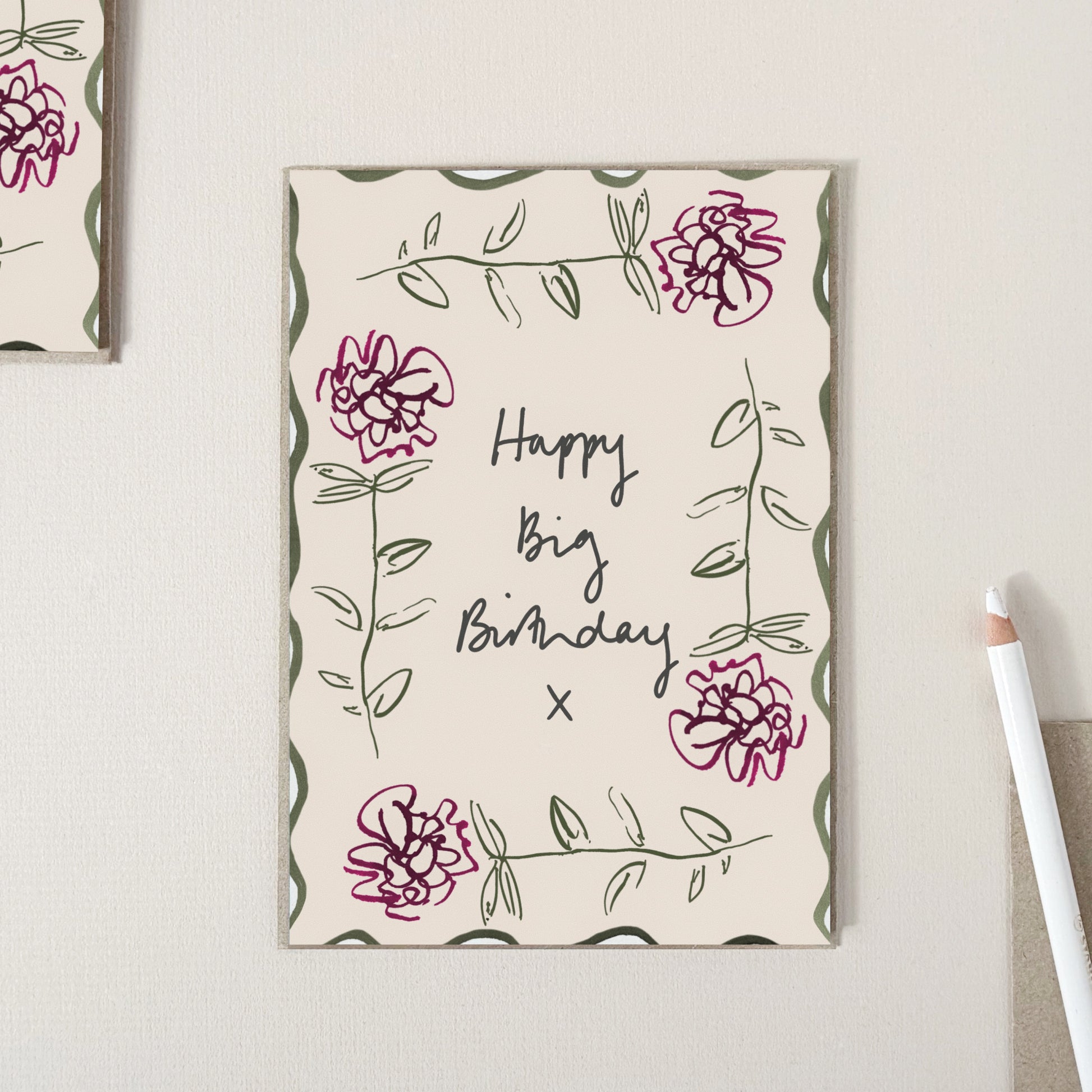 A birthday card with a cream background, featuring hand-drawn green and pink flowers, with the text 'Happy Big Birthday x' in black script. The card is displayed on a light beige textured surface next to a white pencil.