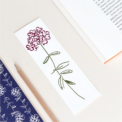 A rectangular bookmark featuring a hand-drawn pink flower with green leaves, placed on a light beige desk alongside a pencil, an open book, and a navy floral notebook.