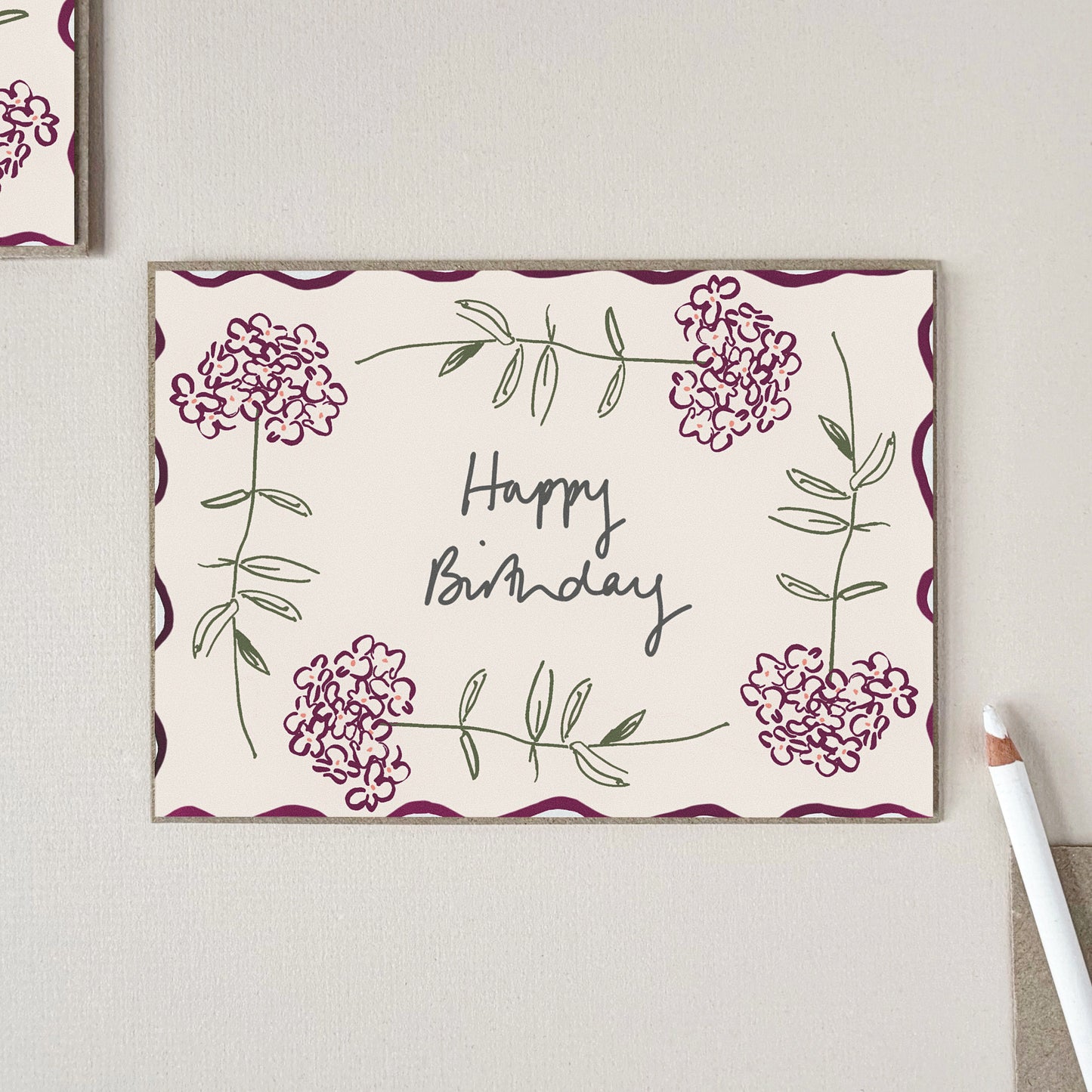 A large birthday card with a warm cream background, featuring four hydrangea stems around 'Happy Birthday' in black script, displayed on a light beige textured surface.