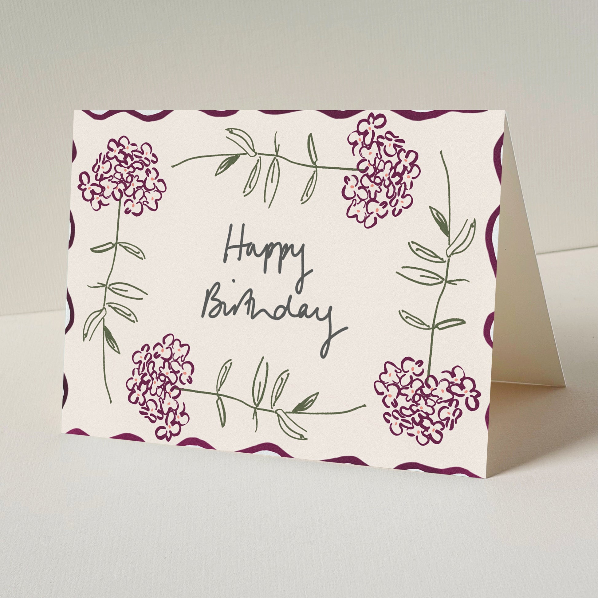 A folded greeting card standing upright on a light surface. The card has a warm cream background with burgundy floral illustrations around the border and the words 'Happy Birthday' written in black script in the center.
