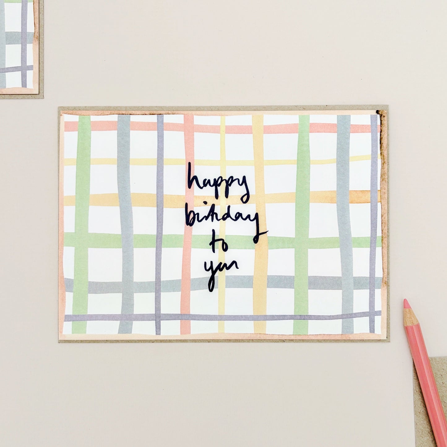 A birthday card with a pastel check pattern in green, yellow, pink, and blue on a white background, featuring the handwritten text 'happy birthday to you' in navy. The card is displayed on a light beige surface next to a pink pencil.
