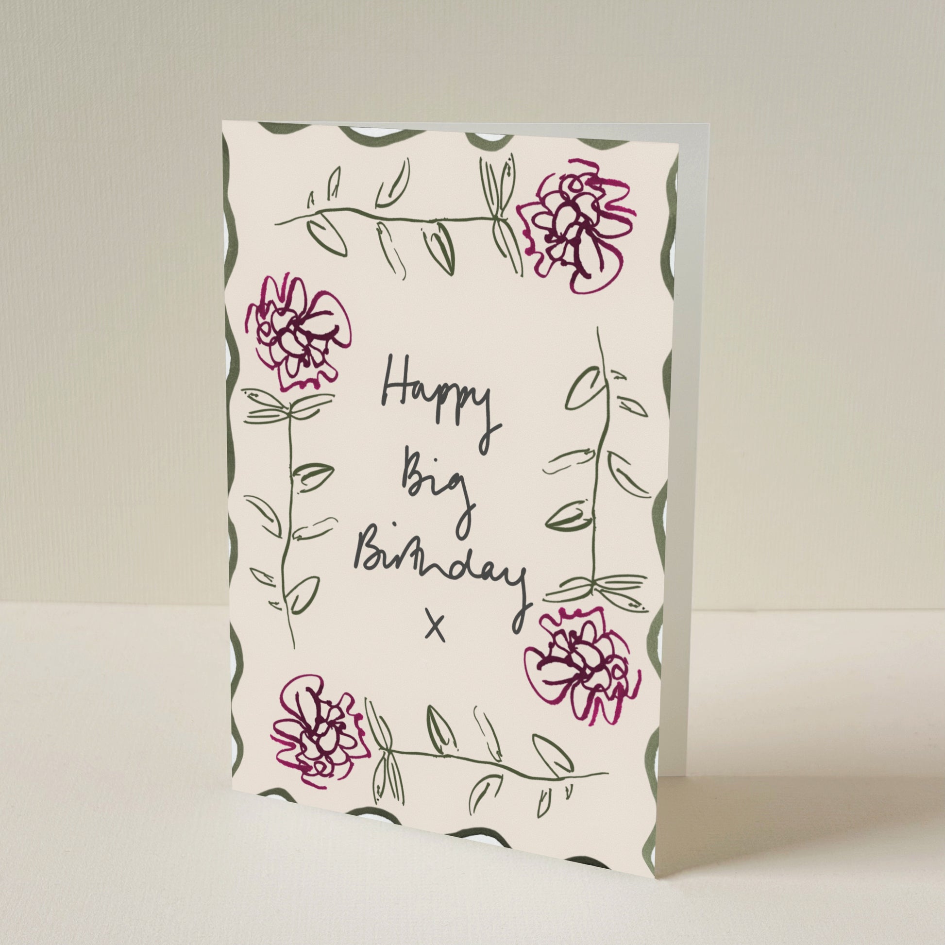 A birthday card with a cream background, featuring hand-drawn green and pink flowers, with the text 'Happy Big Birthday x' in black script. The card is standing upright on a light surface against a neutral light beige background.