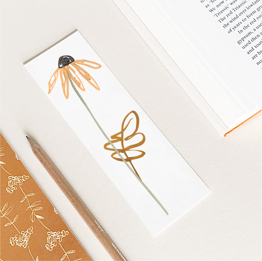 A rectangular bookmark featuring a hand-drawn orange daisy with brown leaf, shown on a light beige desk with a pencil, patterned notebook, and an open book.