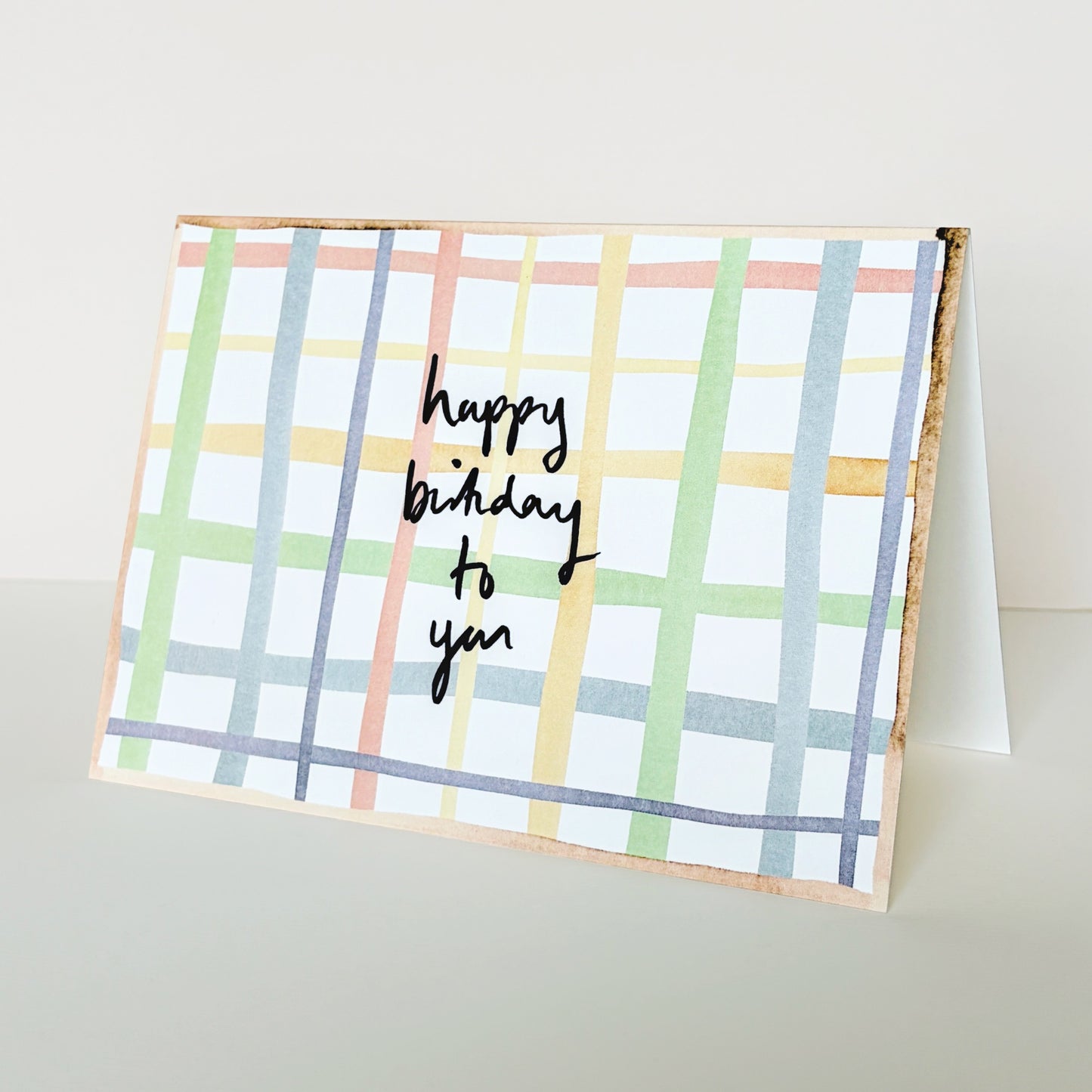 A birthday card with a check  pattern in pastel colours and the handwritten text 'happy birthday to you' in navy ink, displayed on a light beige surface.