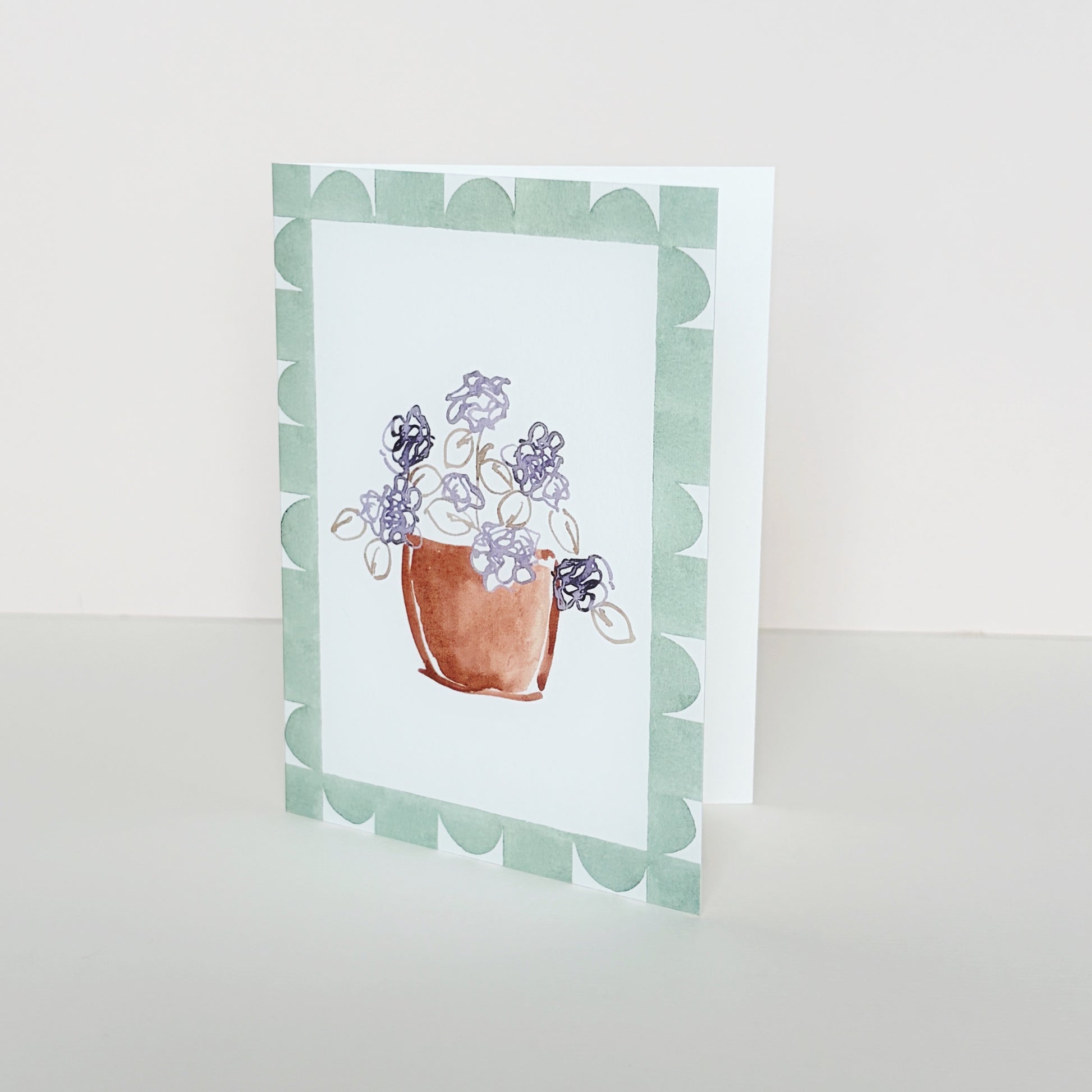 A mini greeting card with a geometric green border and a central ink illustration of a terracotta pot with purple flowers, standing upright on a light beige surface against a cream background.